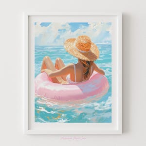 Op de afbeelding: Een ingelijste kunstprint toont een persoon in een strohoed en witte badkleding, ontspannen in een roze opblaasbare ring in de oceaan. Het water is turquoise en de lucht blauw met wolken. De stijl is impressionistisch.