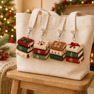 May include: A cream-colored canvas tote bag featuring four hanging crochet keychains. Each keychain consists of a stack of three miniature books in red, green, and cream colors, decorated with holiday motifs like snowflakes, bows, and gingerbread.