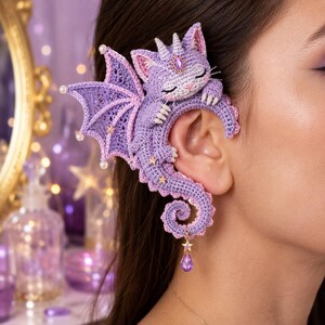 May include: A lavender crochet ear cuff shaped like a sleeping dragon cat with wings, featuring small white pearl accents on the wing tips, gold star embellishments, a purple teardrop gem on the forehead, and a dangling purple crystal charm.
