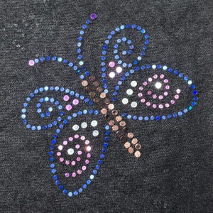 Pu&ograve; includere: Un disegno a farfalla creato con piccoli strass circolari su uno sfondo di tessuto nero. La farfalla presenta un corpo centrale di pietre marroni, con ali delineate in blu e riempite di accenti rosa, bianchi e blu.