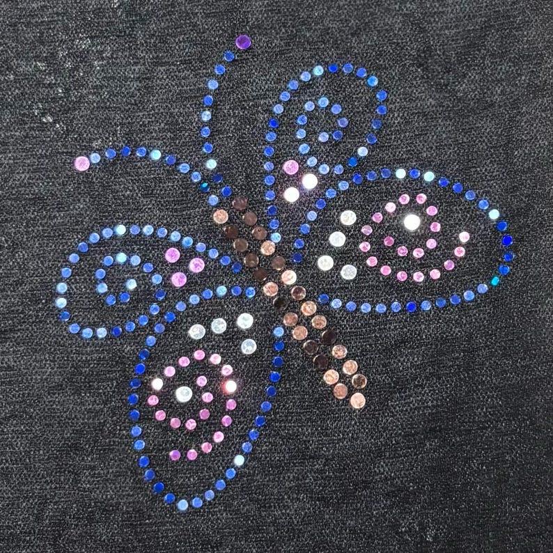 Pu&ograve; includere: Un disegno a farfalla creato con piccoli strass circolari su uno sfondo di tessuto nero. La farfalla presenta un corpo centrale di pietre marroni, con ali delineate in blu e riempite di accenti rosa, bianchi e blu.