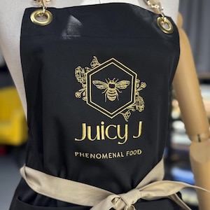 May include: Black apron with gold embroidered bee logo and text "Juicy J Phenomenal Food". The apron has tan straps and a matching waist tie. Gold grommets and hardware are visible.