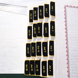 May include: Black Quran tab stickers with gold lettering mark sections of a book. The tabs are labeled with Juz numbers. The text "Quran Tab Stickers" is at the top. The image also includes the text "Durable & Easy to Apply", "Clearly Labeled for Each Juz or Surah", and "Perfect for Marking Quran Sections".