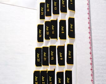 Puede incluir: Pegatinas de índice para Corán en color negro con texto dorado que etiqueta cada Juz del 1 al 28, adheridas a los bordes de un libro. El texto "Quran Tab Stickers" aparece arriba y abajo incluye "Durable & Easy to Apply."