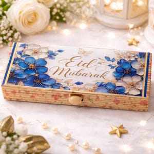 以下が含まれることがあります： 花柄のデザインと金色の「Eid Mubarak」の文字が入った長方形の木箱。青と白の花が特徴で、花、ライト、装飾的な要素に囲まれています。