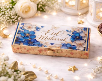 Puede incluir: Una caja rectangular de madera con un diseño floral y la inscripción dorada "Eid Mubarak". La caja presenta flores azules y blancas y está rodeada de flores, luces y elementos decorativos.