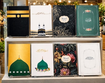 Puede incluir: Ocho libros rectangulares expuestos en un estante de dos niveles. Las portadas presentan diversos diseños, incluyendo la Kaaba, cúpulas verdes, motivos florales y caligrafía árabe. Algunos libros incluyen la palabra Blessed en un marco ovalado.