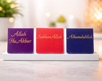 Puede incluir: Tres bloques decorativos cuadrados en morado, rojo y azul, cada uno con escritura dorada. El bloque morado dice "Allah Hu Akbar", el bloque rojo dice "SubhanAllah" y el bloque azul dice "Alhamdulillah". Los bloques se muestran en un soporte blanco.