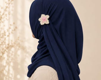 Puede incluir: Un hiyab azul marino liso con un pequeño accesorio de flor de ganchillo de color crema prendido en el lateral. La flor tiene una perla blanca en el centro y la tela del hiyab cae en suaves pliegues verticales a lo largo del costado.
