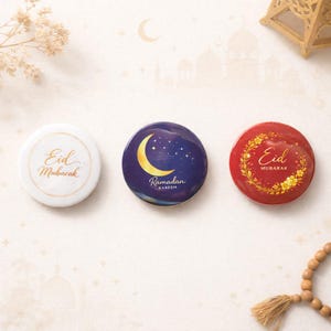 Puede incluir: Tres botones redondos con el tema de Eid Mubarak. Uno es blanco con letras doradas, otro es azul marino con una luna creciente y estrellas, y el tercero es rojo con detalles florales dorados. Los botones son para una celebración.