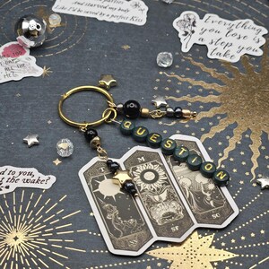 May include: A gold keyring with a charm featuring three tarot cards and the word "QUESTION" spelled out in green beads. The cards have black and white illustrations. The background is dark with gold starburst designs.