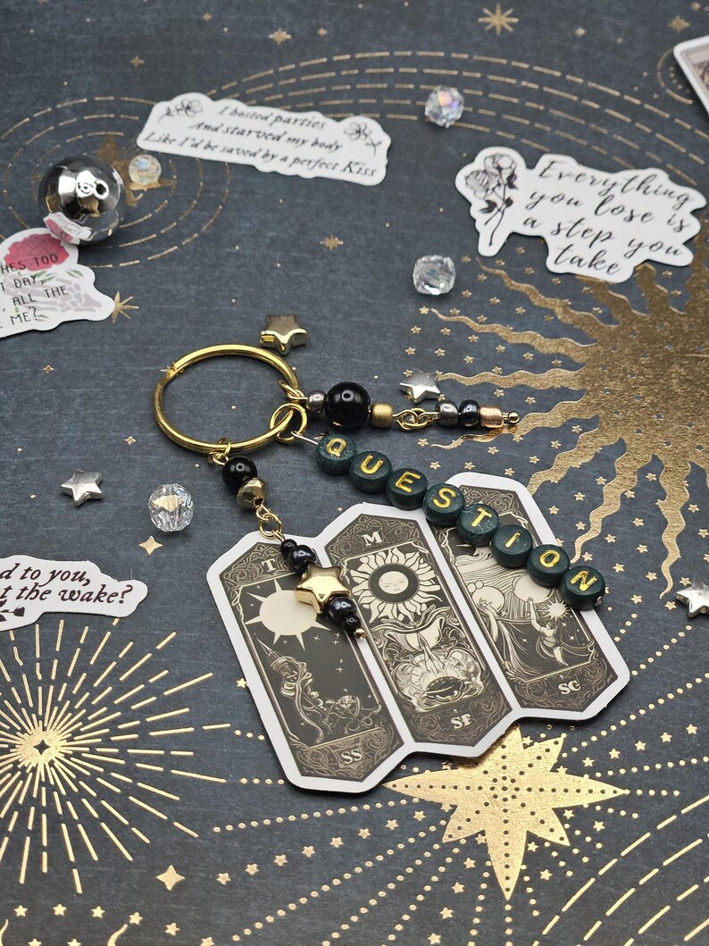 May include: A gold keyring with a charm featuring three tarot cards and the word "QUESTION" spelled out in green beads. The cards have black and white illustrations. The background is dark with gold starburst designs.