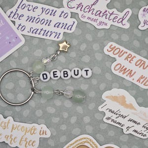 May include: A silver keychain with a beaded charm and the word "DEBUT" spelled out in white letter beads. The charm includes a gold star and green and clear beads. Surrounding the keychain are various stickers with phrases like "love you to the moon and to saturn".