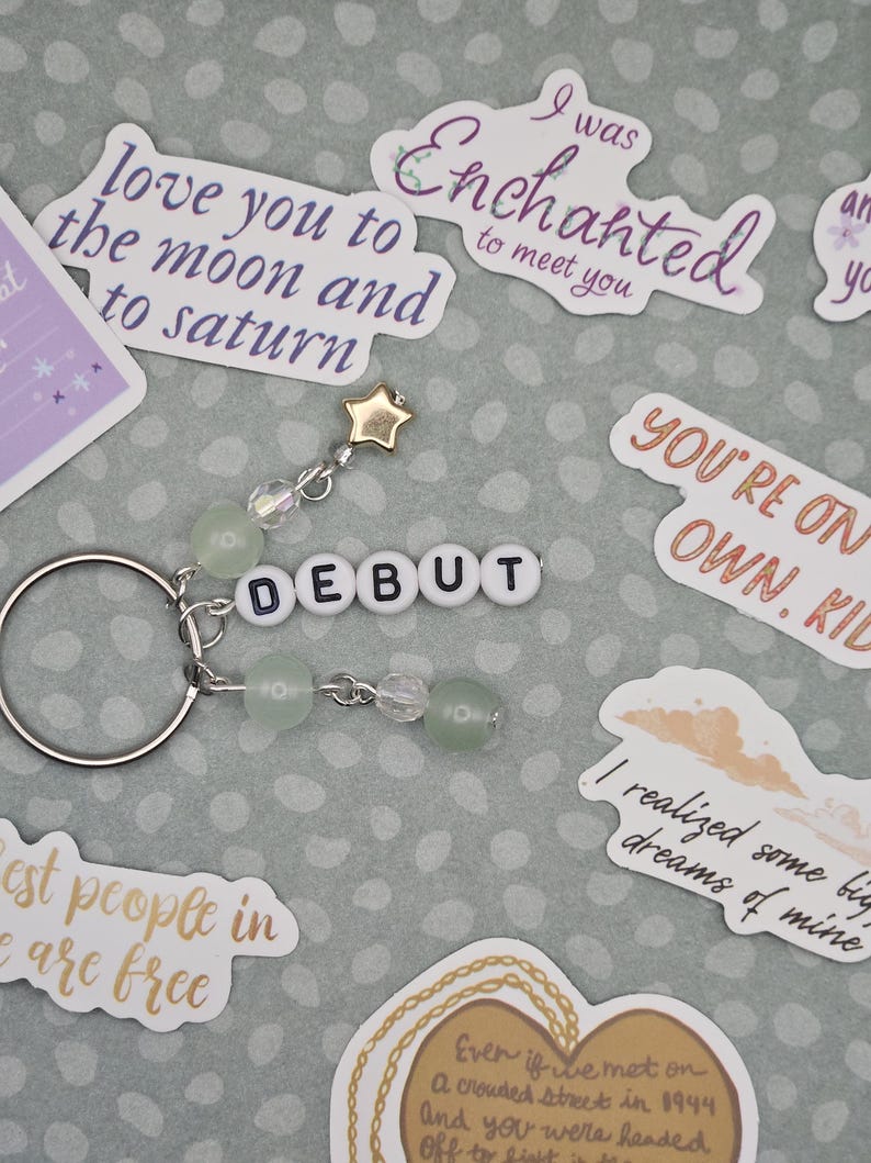 May include: A silver keychain with a beaded charm and the word "DEBUT" spelled out in white letter beads. The charm includes a gold star and green and clear beads. Surrounding the keychain are various stickers with phrases like "love you to the moon and to saturn".