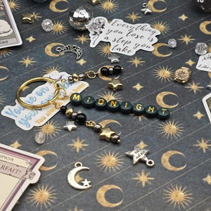 May include: A handmade keychain spelling "MIDNIGHT" in green letter beads, with gold and silver star charms, black beads, and a gold keyring. The background is a dark grey pattern with moons and stars.