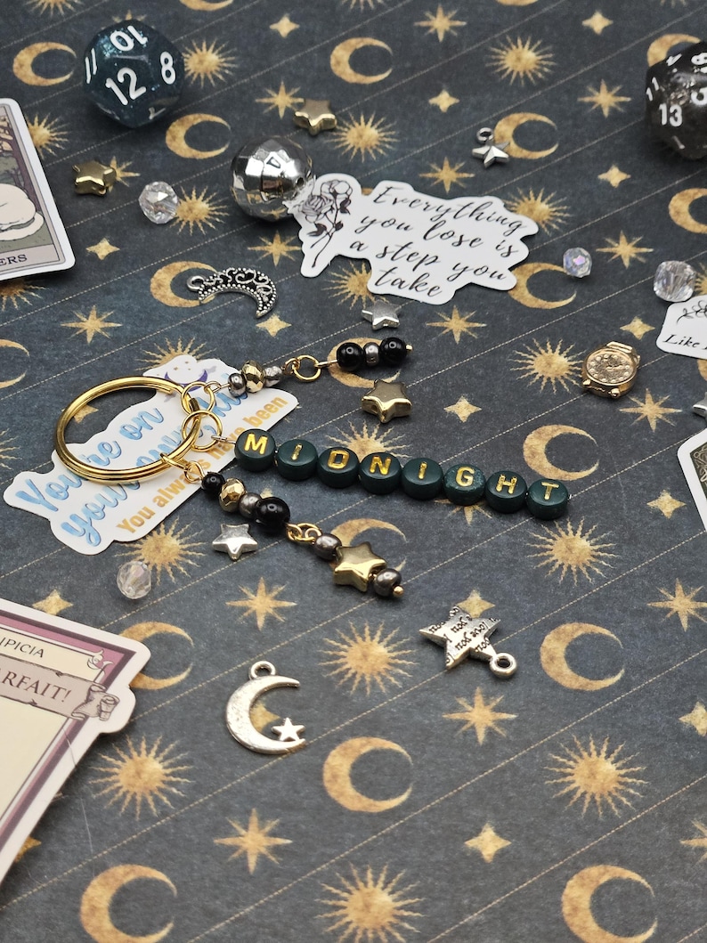 May include: A handmade keychain spelling "MIDNIGHT" in green letter beads, with gold and silver star charms, black beads, and a gold keyring. The background is a dark grey pattern with moons and stars.
