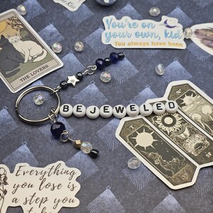 May include: A keychain spelling "BEJEWELED" with white letter beads. It includes a silver star charm, dark blue beads, and a small gold charm. Tarot cards and stickers with text are placed around the keychain. The background is patterned.