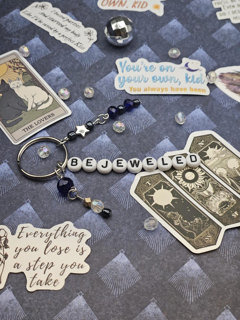 May include: A keychain spelling "BEJEWELED" with white letter beads. It includes a silver star charm, dark blue beads, and a small gold charm. Tarot cards and stickers with text are placed around the keychain. The background is patterned.