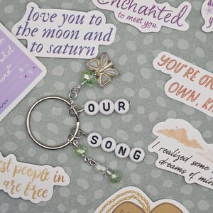 May include: A silver keychain with the words "OUR SONG" spelled out in white letter beads. The keychain features a butterfly charm and green beaded accents. Surrounding the keychain are various stickers with romantic quotes.