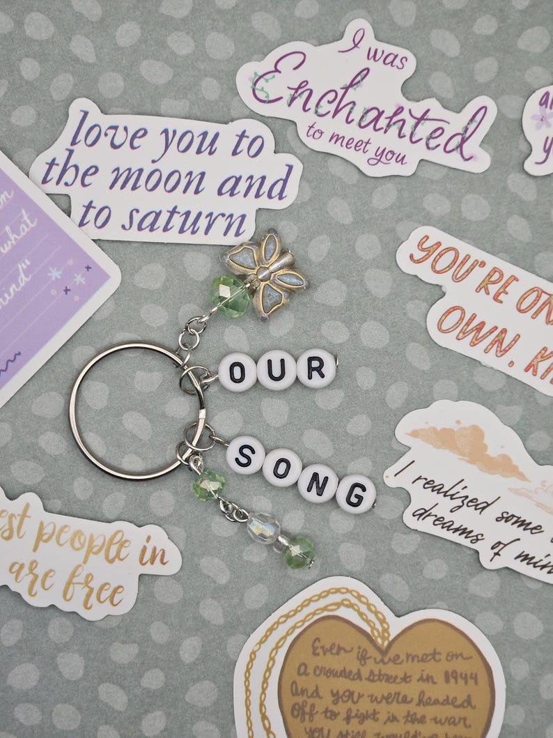May include: A silver keychain with the words "OUR SONG" spelled out in white letter beads. The keychain features a butterfly charm and green beaded accents. Surrounding the keychain are various stickers with romantic quotes.