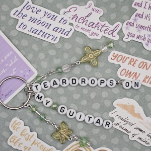 May include: A keychain with the words "TEARDROPS ON MY GUITAR" spelled out in white letter beads. The keychain has green and clear beads, and gold-colored butterfly charms. Surrounding the keychain are stickers with song lyrics.