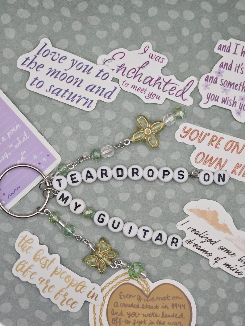 May include: A keychain with the words "TEARDROPS ON MY GUITAR" spelled out in white letter beads. The keychain has green and clear beads, and gold-colored butterfly charms. Surrounding the keychain are stickers with song lyrics.