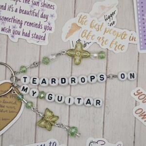May include: A keychain with the words "TEARDROPS ON MY GUITAR" spelled out in white letter beads. The keychain has green and clear beads, and gold-colored charms, including a butterfly and a four-leaf clover.