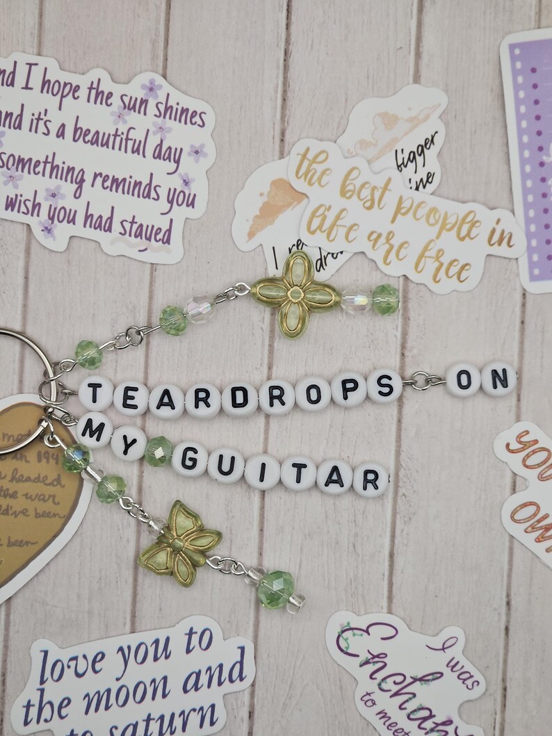May include: A keychain with the words "TEARDROPS ON MY GUITAR" spelled out in white letter beads. The keychain has green and clear beads, and gold-colored charms, including a butterfly and a four-leaf clover.