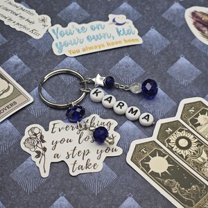 May include: A silver keyring featuring the word "KARMA" spelled out in white letter beads. The keyring has blue crystal beads and a star charm. Stickers with text like "You're on your own, kid" and tarot cards are also visible.