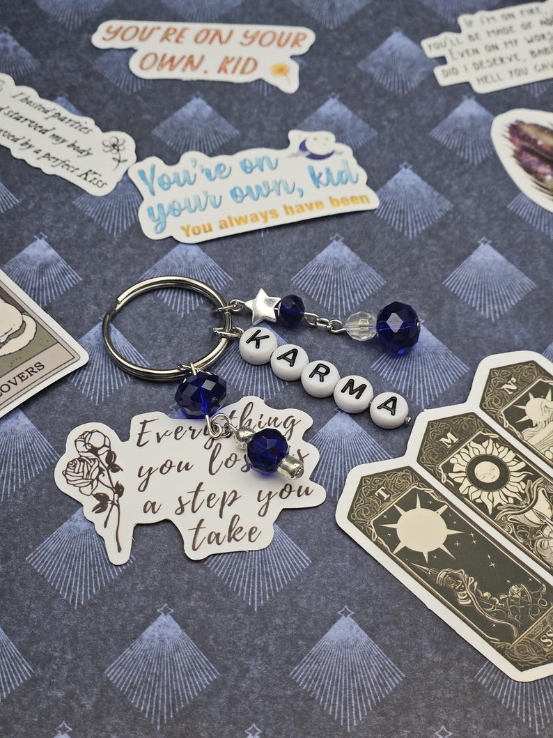 May include: A silver keyring featuring the word "KARMA" spelled out in white letter beads. The keyring has blue crystal beads and a star charm. Stickers with text like "You're on your own, kid" and tarot cards are also visible.