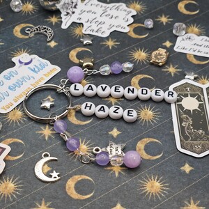 May include: A keychain spelling "LAVENDER HAZE" with white letter beads. It includes purple beads, star charms, and a silver crescent moon charm. The background is a dark fabric patterned with moons and stars.