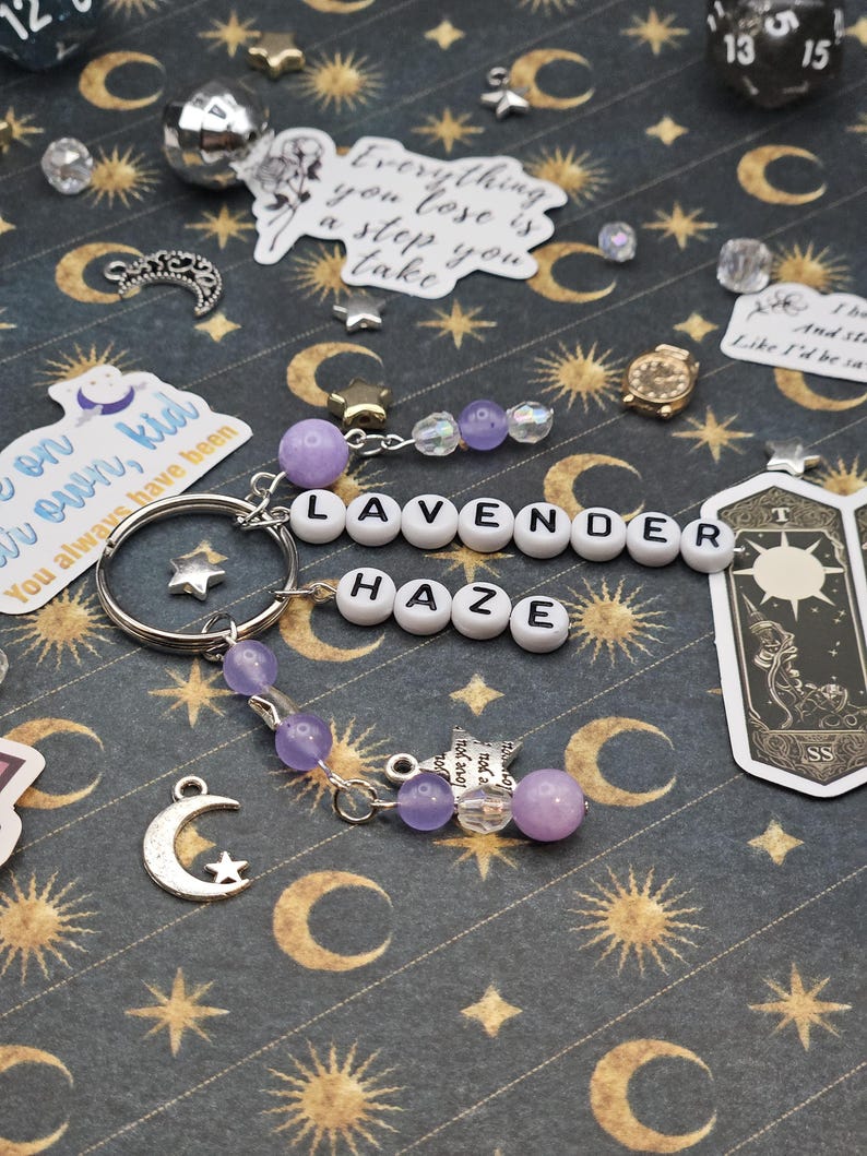 May include: A keychain spelling "LAVENDER HAZE" with white letter beads. It includes purple beads, star charms, and a silver crescent moon charm. The background is a dark fabric patterned with moons and stars.