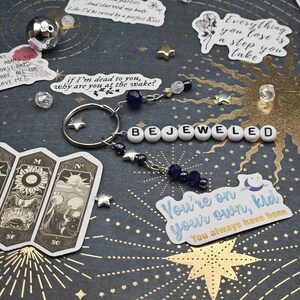 May include: A silver keyring with the word "BEJEWELED" spelled out in white letter beads. The keyring has star charms and dangling beads in shades of blue and clear. Decorative stickers and star-shaped embellishments are scattered around.