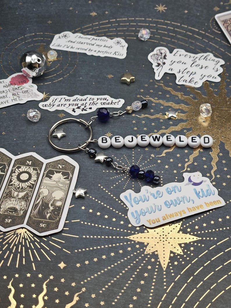 May include: A silver keyring with the word "BEJEWELED" spelled out in white letter beads. The keyring has star charms and dangling beads in shades of blue and clear. Decorative stickers and star-shaped embellishments are scattered around.
