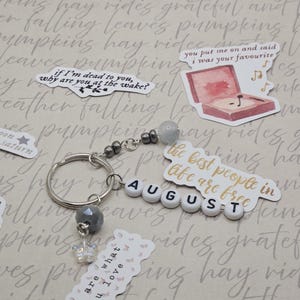 May include: A silver keyring with grey beads and a butterfly charm. The keyring spells "AUGUST" with white letter beads. Decorative stickers with phrases and a record player are scattered around the keyring.
