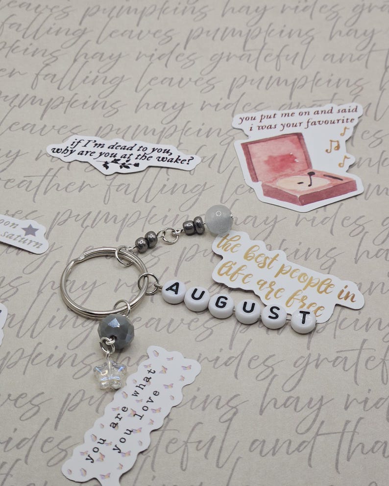 May include: A silver keyring with grey beads and a butterfly charm. The keyring spells "AUGUST" with white letter beads. Decorative stickers with phrases and a record player are scattered around the keyring.