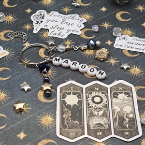 May include: A keyring spelling "MAROON" in white letter beads, with star charms and a small gold clock charm. The background is a dark patterned surface with celestial motifs and stickers. The image also includes a dice and tarot card stickers.