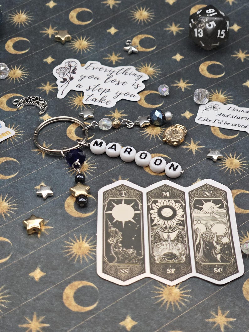 May include: A keyring spelling "MAROON" in white letter beads, with star charms and a small gold clock charm. The background is a dark patterned surface with celestial motifs and stickers. The image also includes a dice and tarot card stickers.