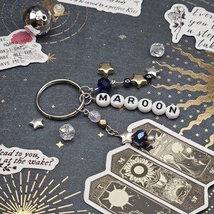 May include: A keychain spelling "MAROON" in white letter beads, with star charms and a dark blue bead. Attached are three tarot card stickers. The background is dark with gold star and sunburst patterns.