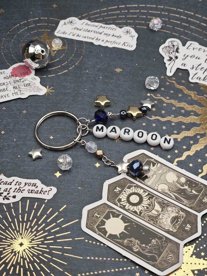 May include: A keychain spelling "MAROON" in white letter beads, with star charms and a dark blue bead. Attached are three tarot card stickers. The background is dark with gold star and sunburst patterns.