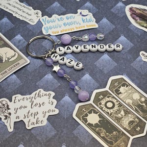 May include: A silver keyring with beaded charms spelling "LAVENDER HAZE." The charms feature purple beads and star accents. Stickers with text, including "You're on your own, kid" and tarot cards are scattered around the keyring.