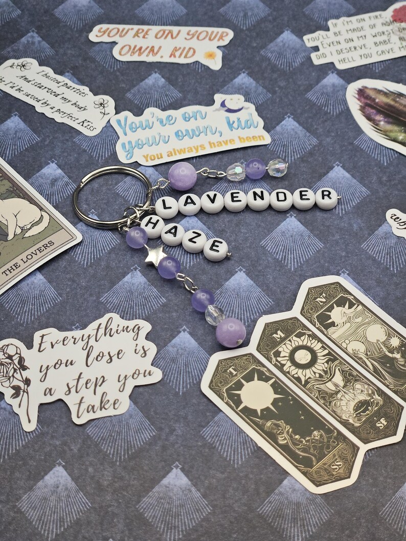 May include: A silver keyring with beaded charms spelling "LAVENDER HAZE." The charms feature purple beads and star accents. Stickers with text, including "You're on your own, kid" and tarot cards are scattered around the keyring.