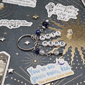 May include: A silver keyring featuring the words "ANTI HERO" spelled out in white letter beads. The keyring is decorated with blue and clear beads. Stickers with text and star-shaped embellishments are scattered around the keyring.