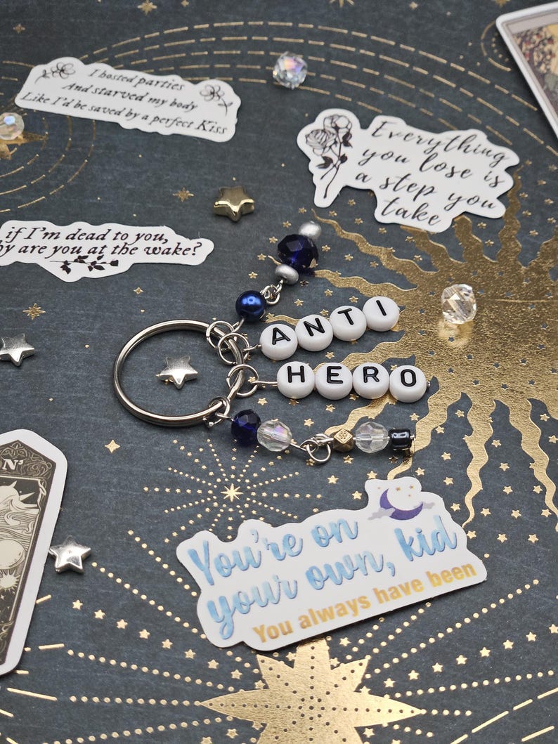 May include: A silver keyring featuring the words "ANTI HERO" spelled out in white letter beads. The keyring is decorated with blue and clear beads. Stickers with text and star-shaped embellishments are scattered around the keyring.
