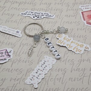 May include: A silver keyring with the name "BETTY" spelled out in white letter beads. The keyring has decorative beads and charms, and is surrounded by stickers with quotes and illustrations.
