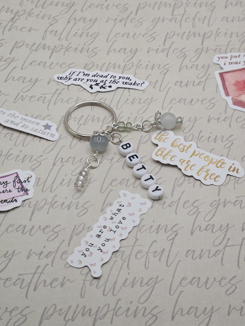 May include: A silver keyring with the name "BETTY" spelled out in white letter beads. The keyring has decorative beads and charms, and is surrounded by stickers with quotes and illustrations.