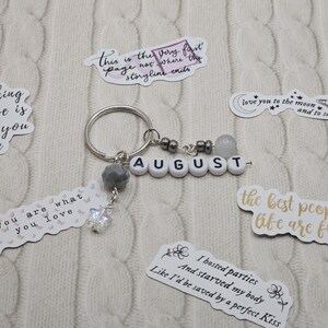 May include: A silver keyring with the name "AUGUST" spelled out in white letter beads. The keyring has grey and clear bead accents and a small, dangling charm. Several stickers with quotes and phrases surround the keyring.