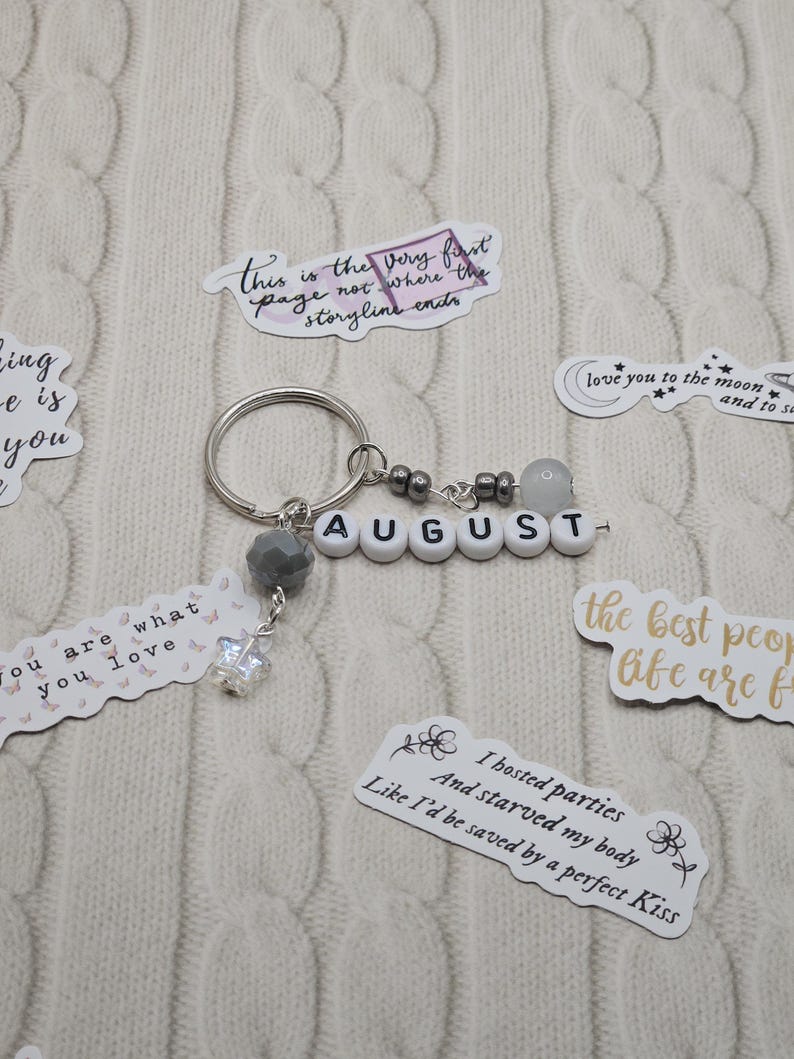 May include: A silver keyring with the name "AUGUST" spelled out in white letter beads. The keyring has grey and clear bead accents and a small, dangling charm. Several stickers with quotes and phrases surround the keyring.