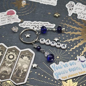 May include: A silver keyring with the word "KARMA" spelled out in white letter beads. The keyring features blue and clear crystal beads and star charms. The background is a dark blue surface with gold star and celestial designs, and stickers with text.