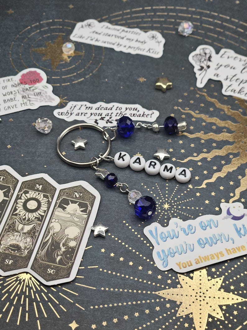 May include: A silver keyring with the word "KARMA" spelled out in white letter beads. The keyring features blue and clear crystal beads and star charms. The background is a dark blue surface with gold star and celestial designs, and stickers with text.